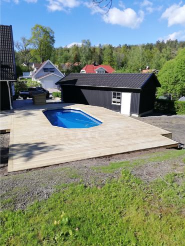 Trädäck med en liten, rektangulär pool på en bakgård, omgiven av gräs och träd, med svarta byggnader i närheten under en blå himmel.