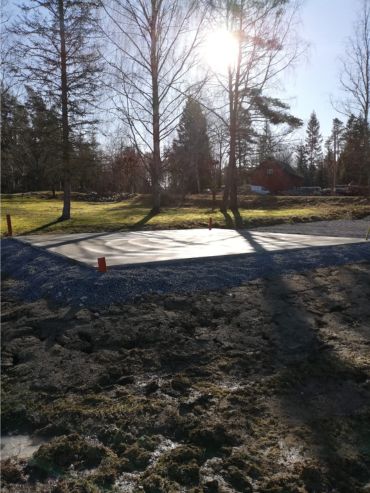 En betongplatta ligger i ett gräsområde med träd. Solen syns bakom träden och kastar långa skuggor över marken.