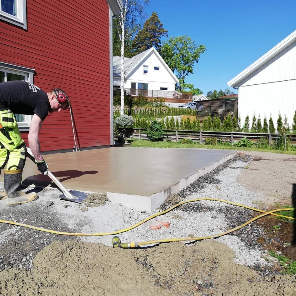 Iförd säkerhetsutrustning planerar en person sakkunnigt våt betong på en uteplats bredvid ett rött hus, med två andra synliga i bakgrunden. Ett sandområde och trädgårdsslang föreslår nyligen grävarbete. Det är en solig dag perfekt för sådana projekt.