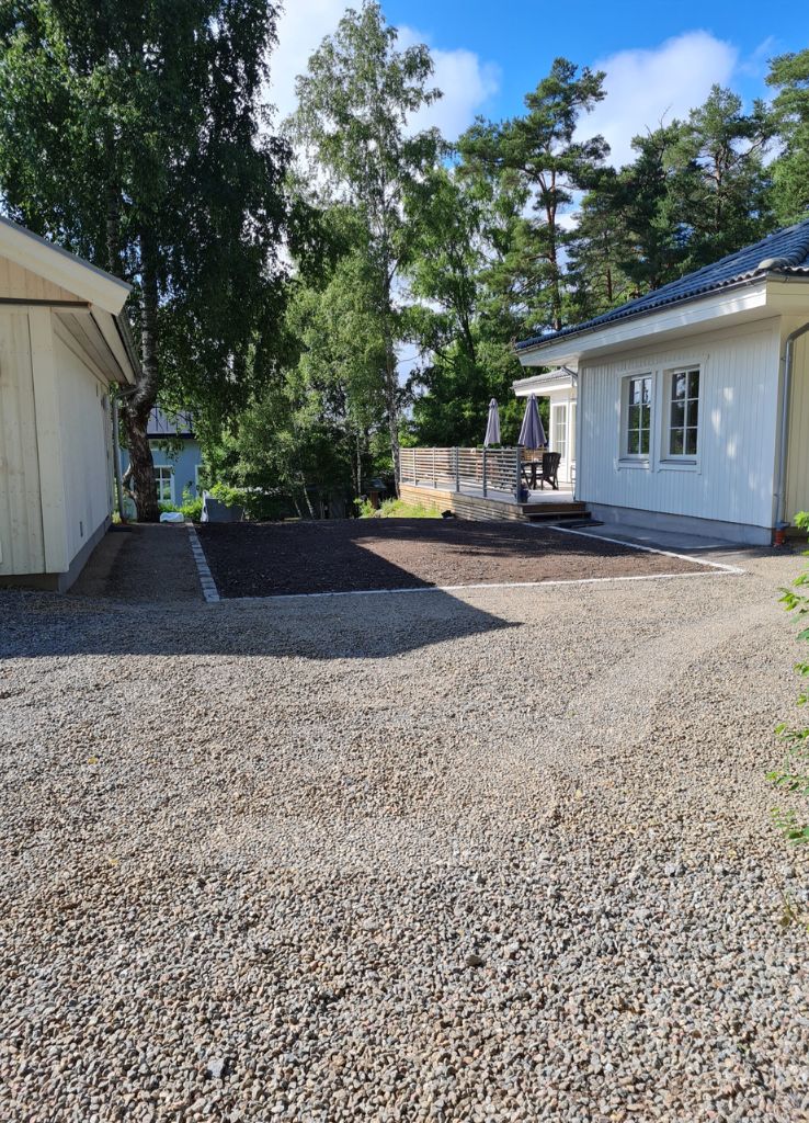 Grusuppfart som leder till ett vitt hus med sluttande tak och en angränsande öppen gård. Träd och ett fristående garage syns i bakgrunden.