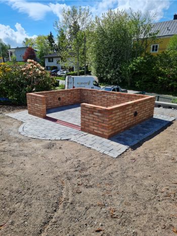 En delvis konstruerad tegelkonstruktion vilar på en stenlagd yta bland smutsen och träden, med en vit skåpbil i bakgrunden. Den här scenen verkar redo för ytterligare markarbete, och antyder framtida landskapsarkitektur eller grävarbete för att sömlöst smälta in i sin naturliga omgivning.