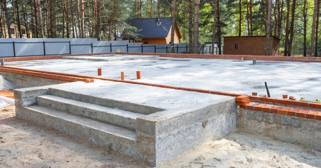 En betongfundament med tegelkant och flera rörledningar ligger i ett skogsområde, med betongtrappor som leder upp till det. Ett trästaket, ett litet hus och ett skjul syns i bakgrunden bland träden.