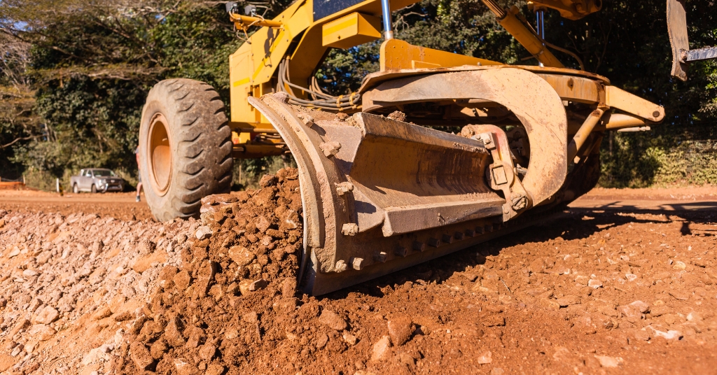 Närbild på väghyvel som skrapar och jämnar ut grus och jord på en väg, med träd i bakgrunden.