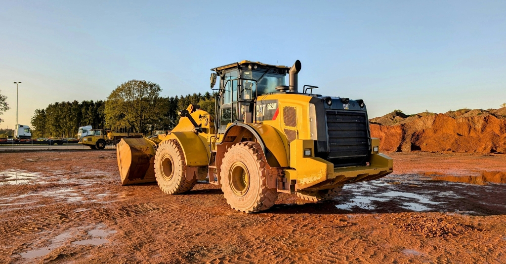 Gul hjullastare på en lerig byggarbetsplats med jordhögar och träd i bakgrunden.