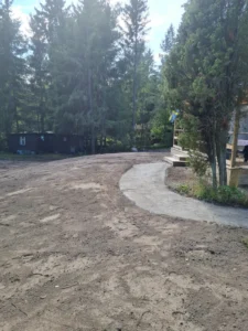 En grusstig kröker sig genom ett område med glest gräs, vilket leder till en skog av höga vintergröna träd. En delvy av en byggnad med en veranda är till höger.